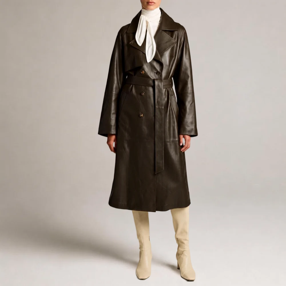 A model wearing a dark brown double-breasted leather trench coat with a matching belt, styled with a white neck scarf, cream turtleneck, and beige boots, shot against a neutral light gray studio background.