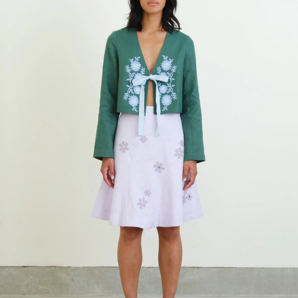 A model wearing a green long-sleeve linen cropped jacket with white floral embroidery and a light blue tie, paired with a light purple skirt with purple floral prints, set against a plain off-white wall.