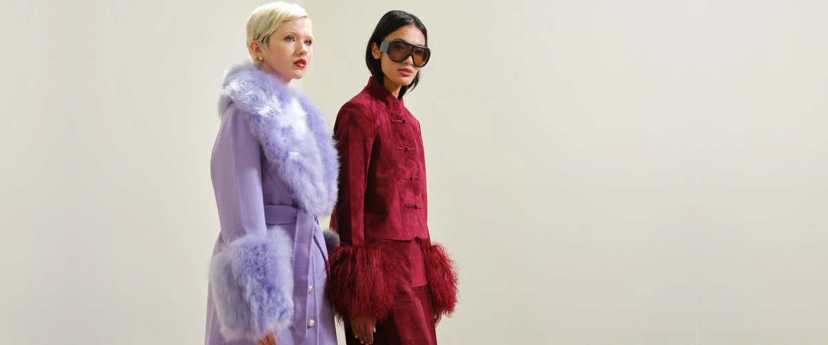 Two models standing against a plain light gray studio background. The model on the left wears a lavender coat with fluffy pale lavender fur trim. The model on the right is wearing oversize sunglasses and a burgundy suede jacket with fur trim sleeves and a matching skirt.