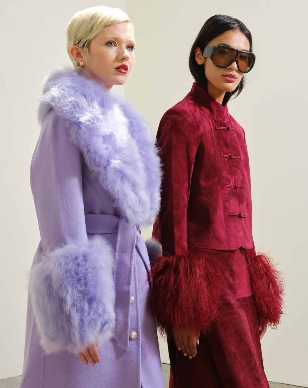 Two models standing against a plain light gray studio background. The model on the left wears a lavender coat with fluffy pale lavender fur trim. The model on the right is wearing oversize sunglasses and a burgundy suede jacket with fur trim sleeves and a matching skirt.