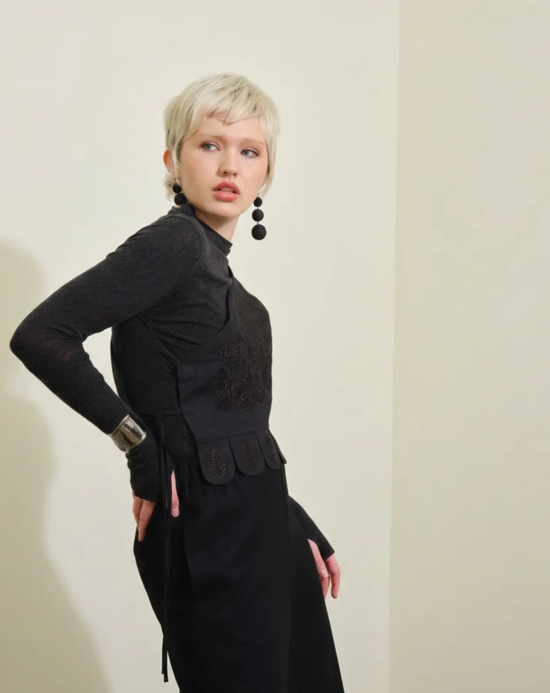 A model with short blonde hair wearing a dark grey long-sleeve cashmere top, embroidered linen black asymmetrical vest,and black dangling earrings, standing against a plain beige wall with a soft shadow.