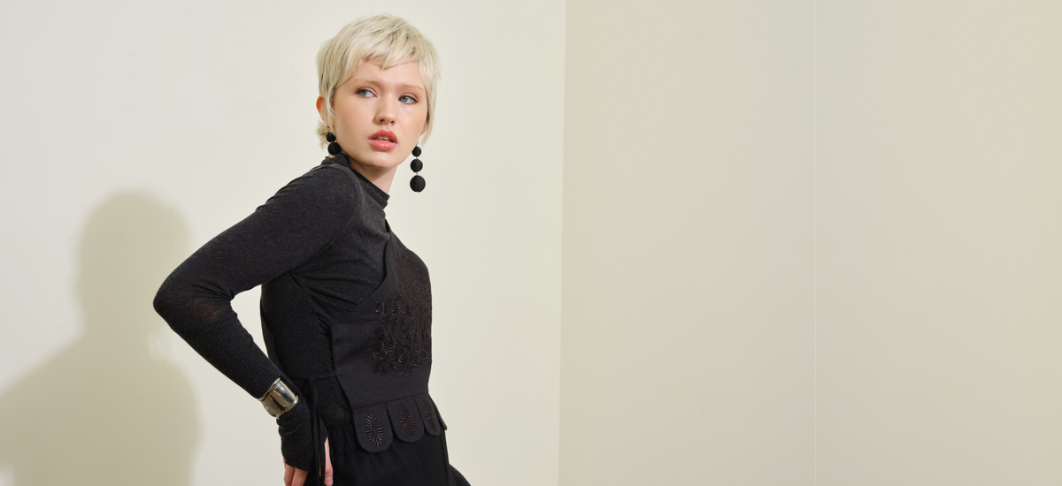 A model with short blonde hair wearing a dark grey long-sleeve cashmere top, embroidered linen black asymmetrical vest,and black dangling earrings, standing against a plain beige wall with a soft shadow.
